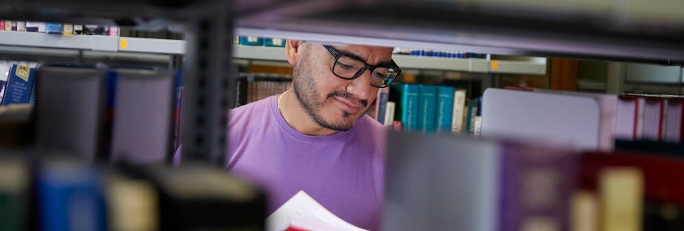 A man with a beard and glasses reading a book in a library.